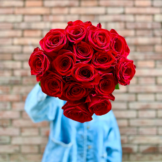 Bouquet of red roses held by a person wearing a blue denim jacket against a brick wall.